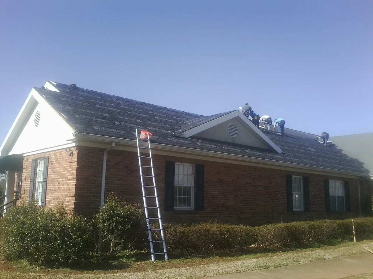 Crew tearing shingles off a brick ranch roof with a ladder set — Metal Roof Repair work by our crew in Bradyville