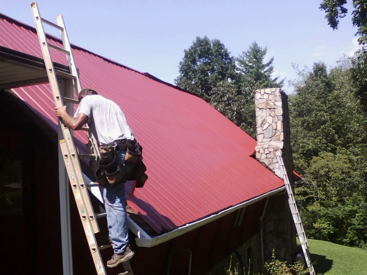 Roofer on a ladder beside a red metal roof and stone chimney — Soffit & Fascia Repair by Roofing Brothers in Bradyville